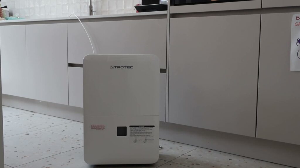 Déshumidificateur Trotec TTK 127 E devant les meubles de cuisine, tuyau d’évacuation blanc relié à l’évier