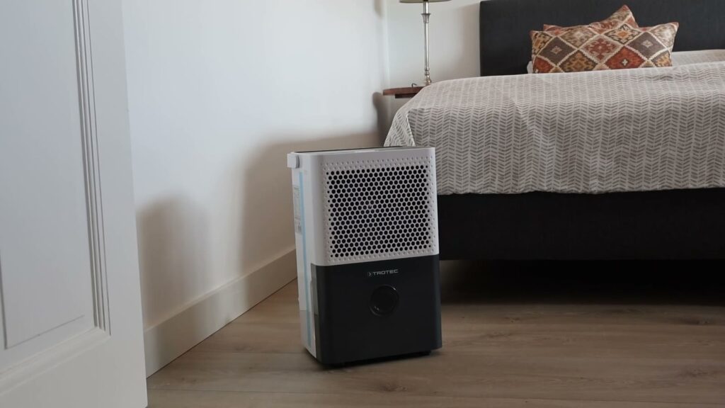 Déshumidificateur Trotec TTK 33 E posé sur le parquet au pied d’un lit, dans une chambre